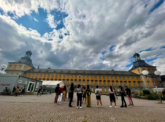 Eine Wissenschaftlerin und ein Wissenschaftler arbeiten hinter einer Glasfassade und mischen Chemikalien mit Großgeräten.